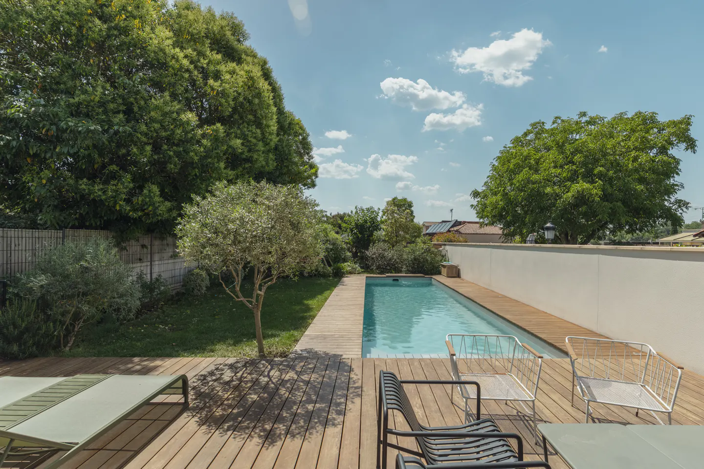 A rectangular swimming pool set in a wooden deck with modern outdoor chairs, a green lawn, and lush trees under a blue sky.