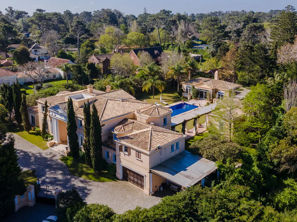 Aerial view of a large, tan mansion with a tile roof, surrounded by green trees and a blue swimming pool.