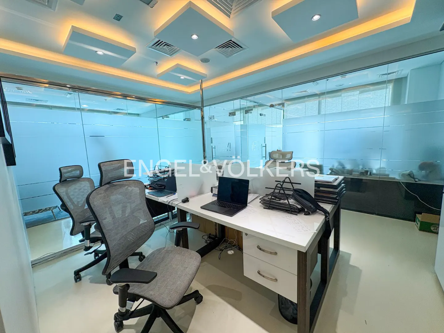 Bright office space with glass walls, desks, and gray chairs. Laptops and office supplies are on the desks. The ceiling has recessed lighting.