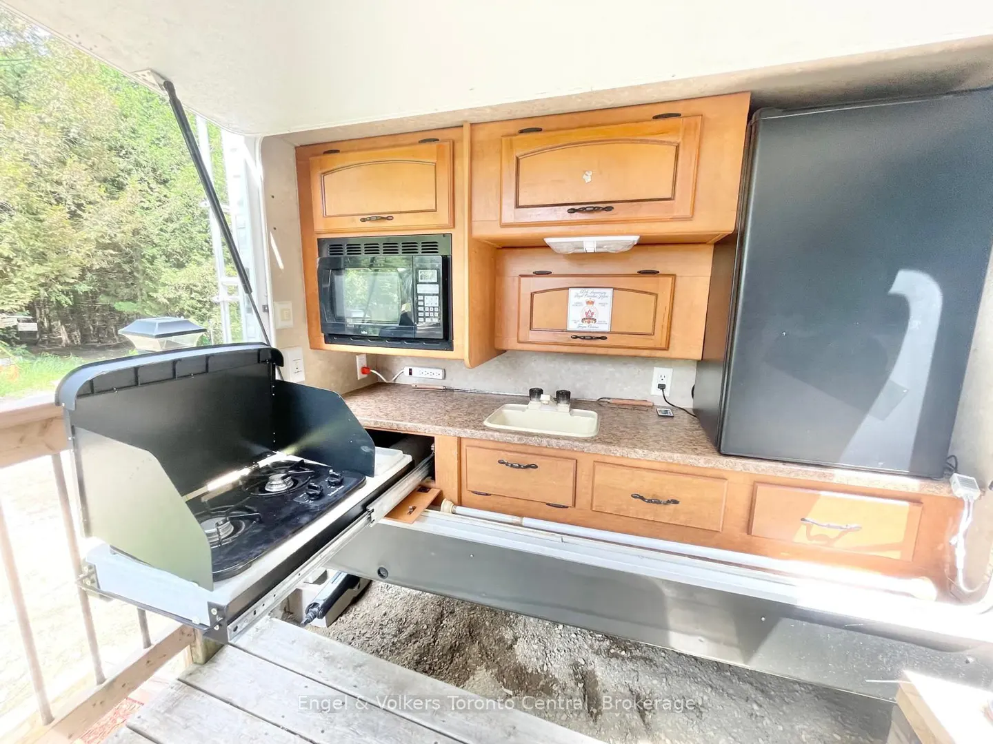 RV kitchen with wood cabinets, microwave, sink, and black refrigerator. A pull-out stove is on a wooden deck with trees in the background.