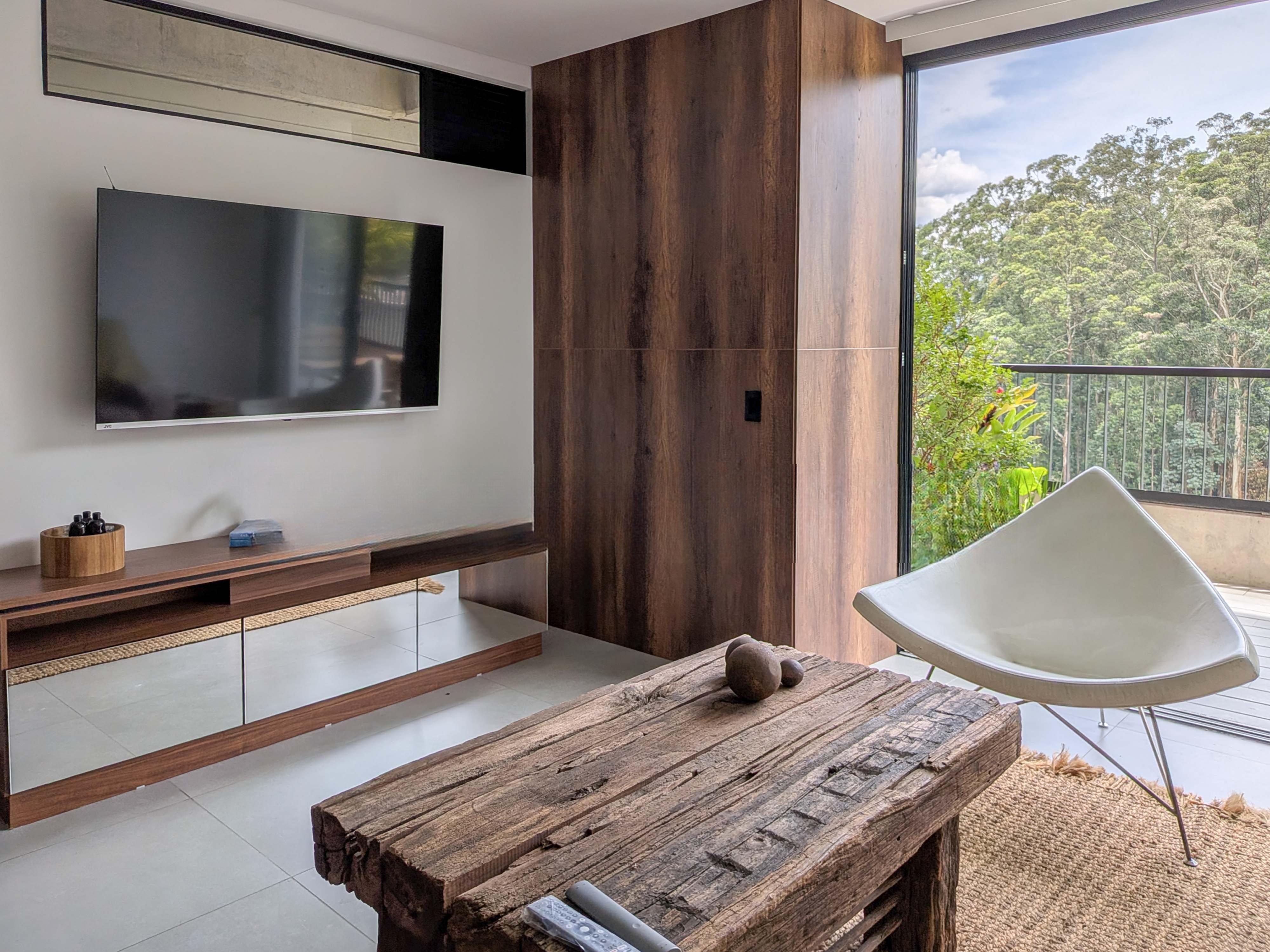 Living room with TV, wood cabinet, and rustic coffee table. A white chair faces a window with a view of lush greenery.