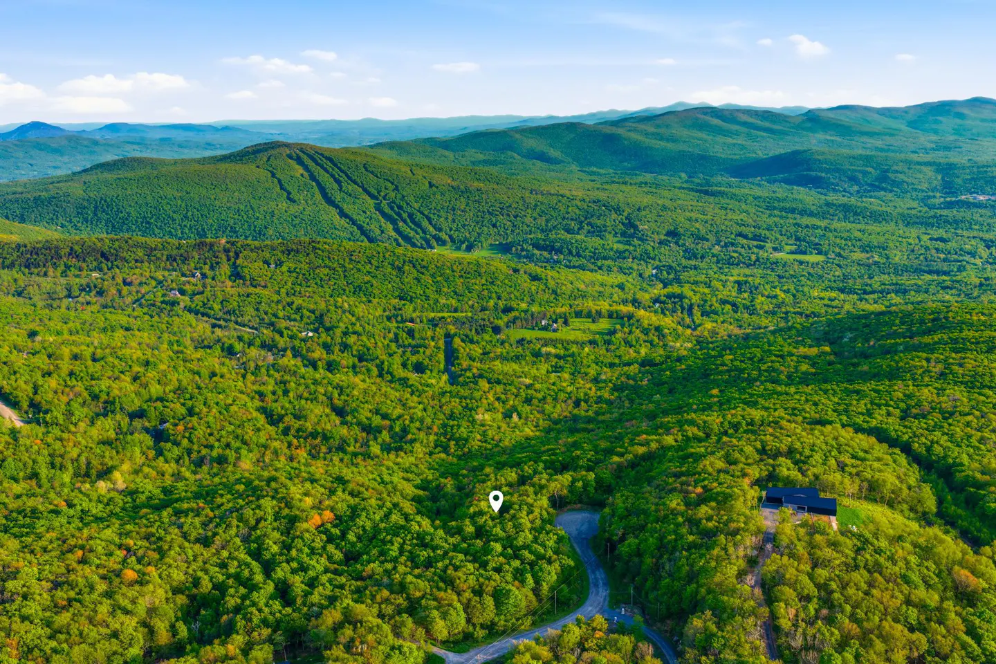 Aerial view of a lush green mountain landscape with a winding road, a modern home, and a map pin marking a property lot.