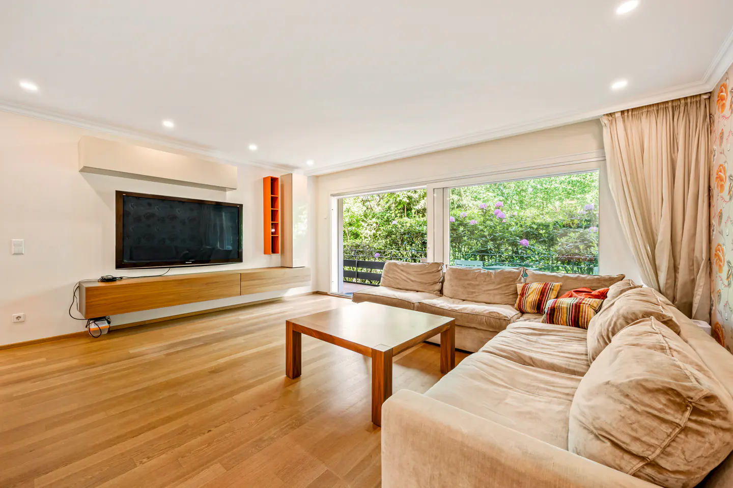 Living room with hardwood floors, a large beige sectional sofa, and a wooden coffee table. A TV is mounted above a wooden console. Large windows overlook a green garden.