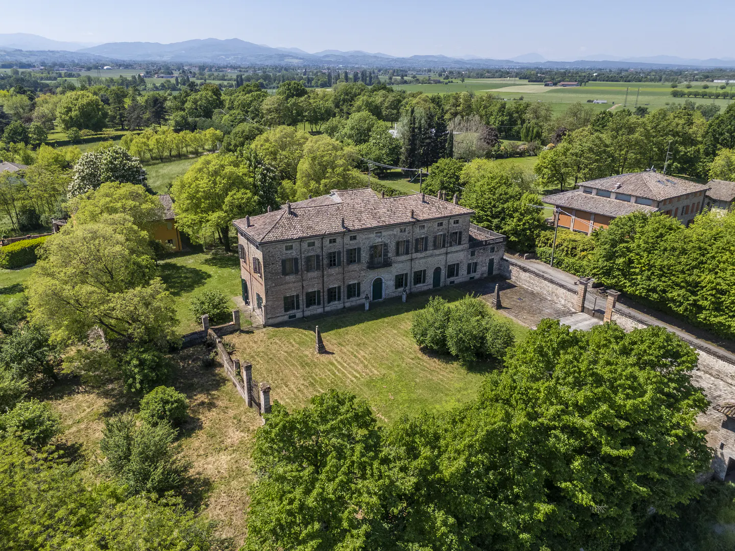 Aerial view of a large, two-story brick house with a red tile roof, surrounded by green trees and lawn, with mountains in the distance.