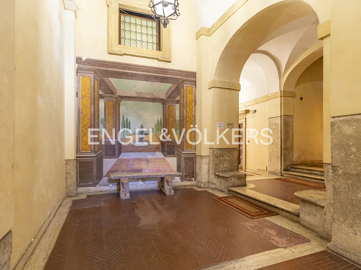 Interior view of a hallway with arches, a mural, and a stone bench. The walls are yellow, and the floor is red brick. The Engel & Völkers logo is visible.