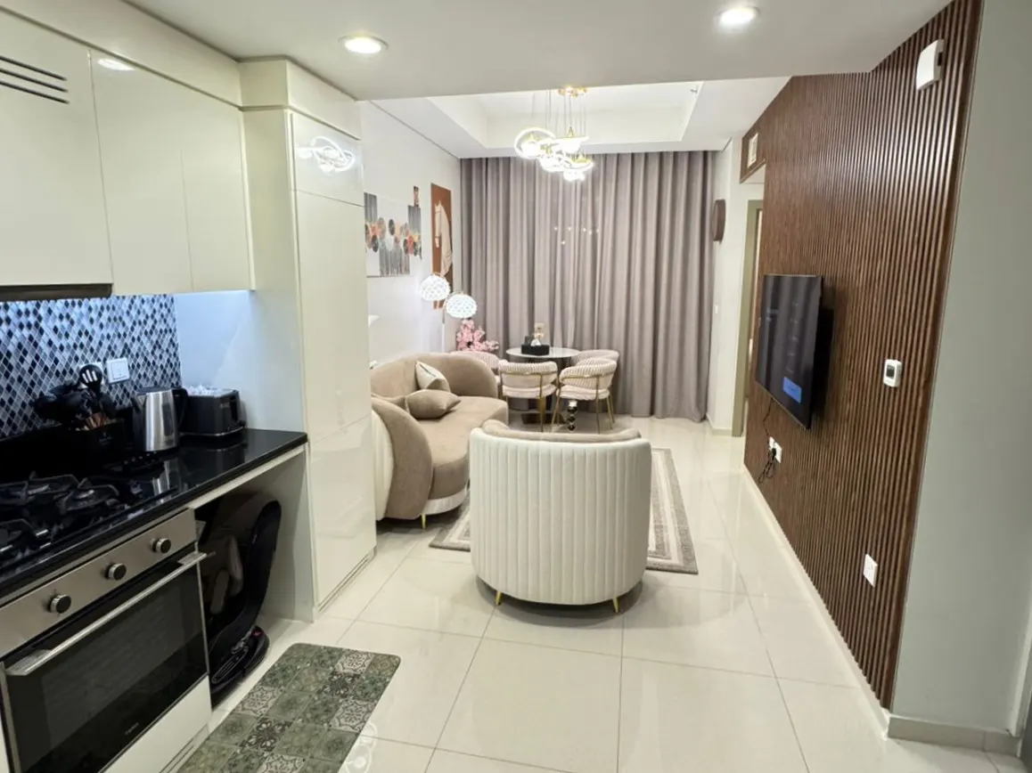 A modern apartment interior with a kitchen, living, and dining area. Beige sofa, white chair, and a TV on a wood-paneled wall.