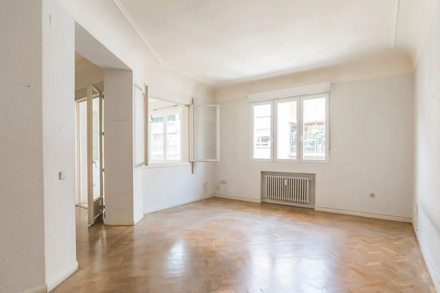 Bright, empty room with parquet floors, white walls, and three windows. A radiator sits beneath the windows. A doorway is visible on the left.