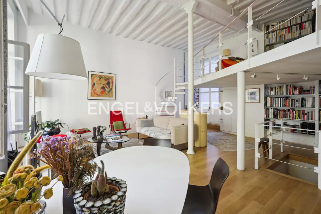 Bright, modern loft interior with white walls, a spiral staircase, and a mezzanine level with bookshelves. A round white table is in the foreground.