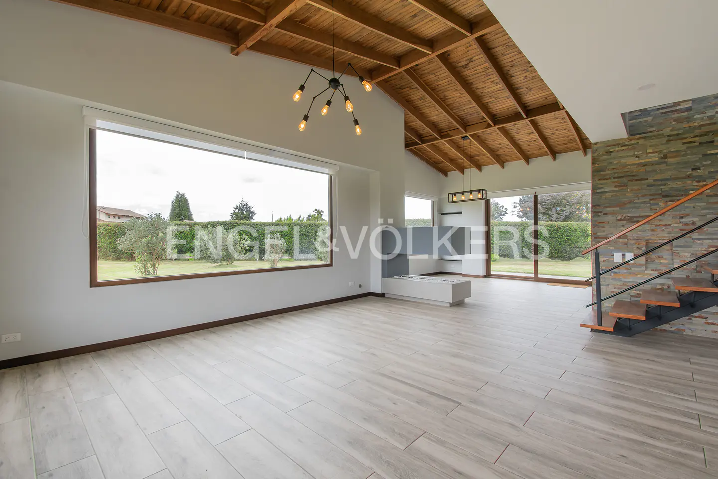 Bright, open living room with wood-beamed ceiling, large windows, and stone accent wall with stairs.