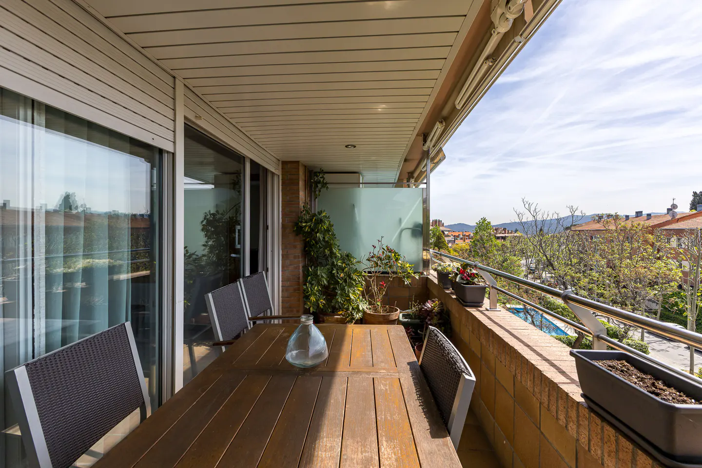 A balcony with a wooden table, chairs, and plants overlooking a cityscape on a sunny day.