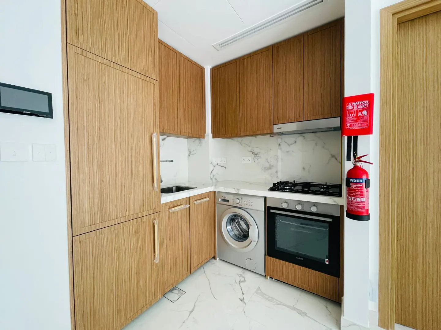 A compact kitchen with wood cabinets, marble countertops, and stainless steel appliances. A red fire extinguisher is mounted on the wall.