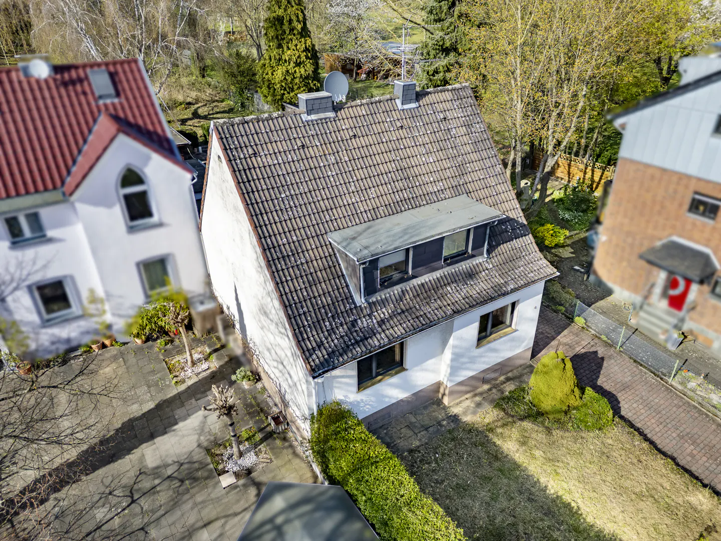Aerial view of a white house with a brown tiled roof, dormer window, and green lawn. Other houses and trees are in the background.