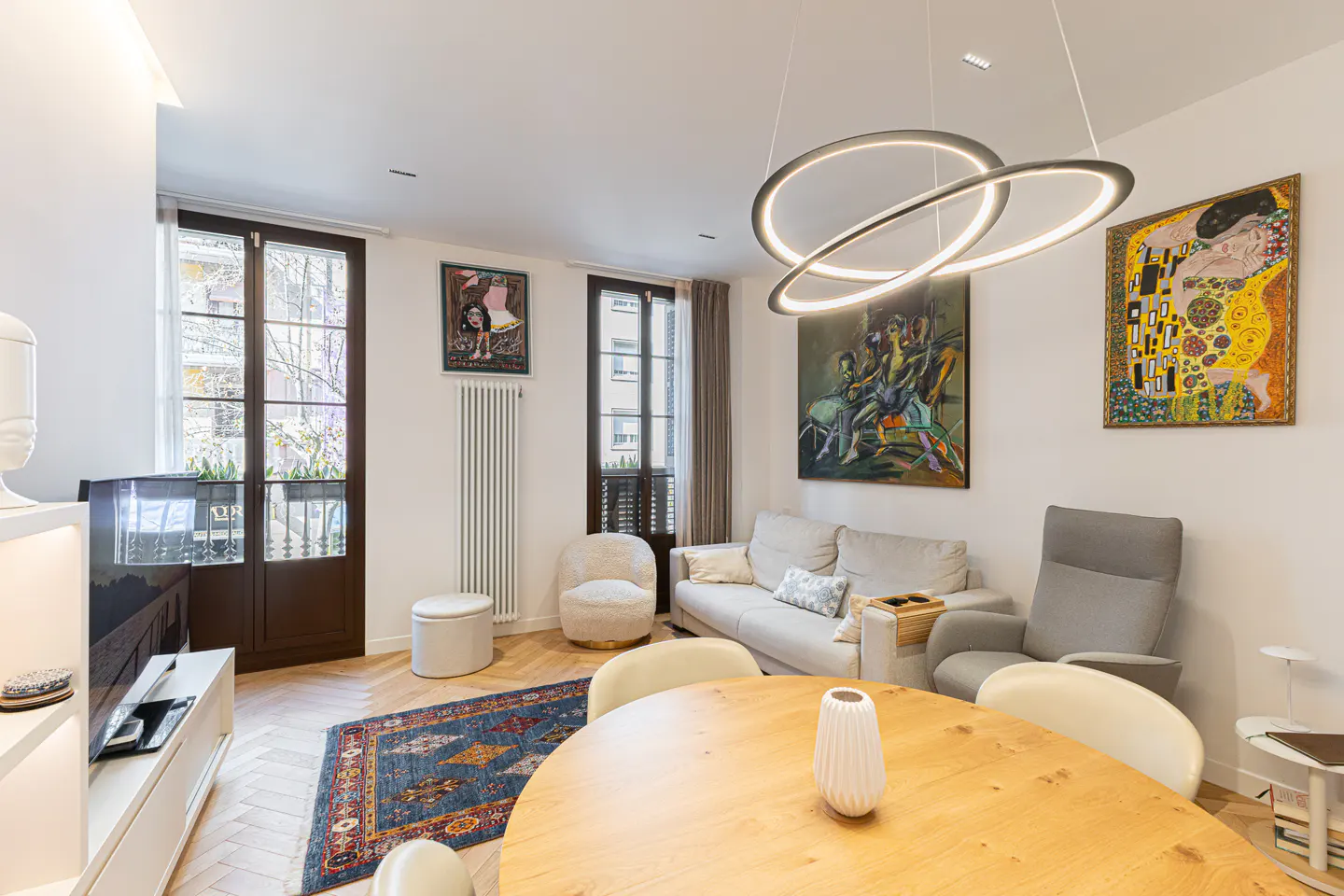 Modern living room with a round wooden dining table, light gray sofa, armchair, and a unique circular LED pendant light.