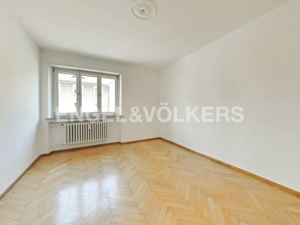 Empty room with herringbone wood floor, white walls, radiator under window, and Engel & Völkers logo on the wall.