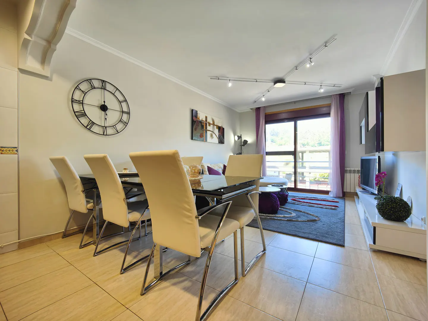 Living room with dining area. A black glass table with cream chairs sits near a large clock. A balcony with purple curtains is in the background.
