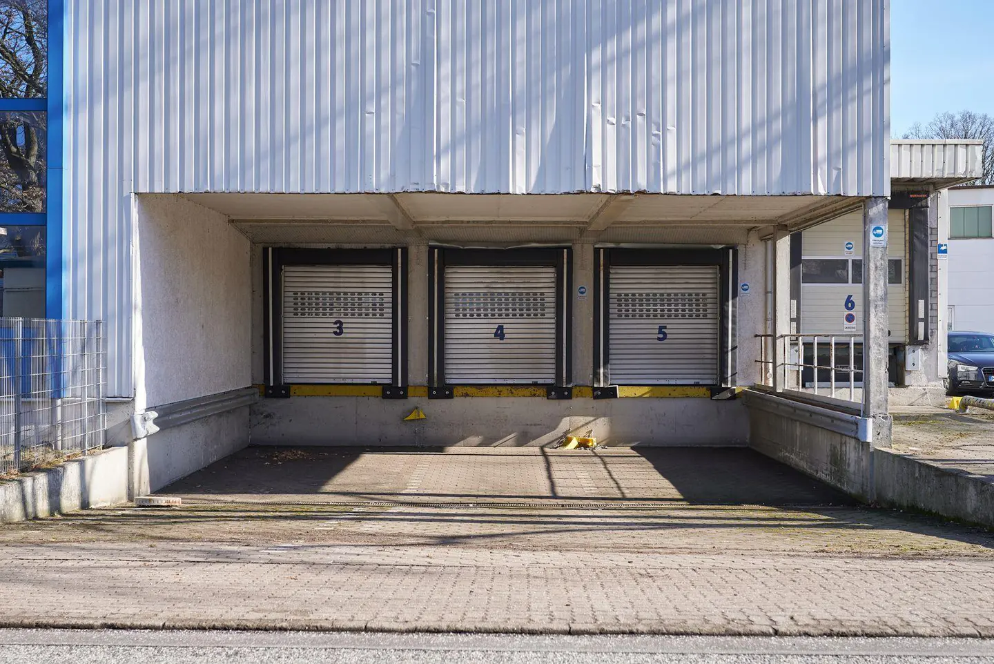 Exterior view of a warehouse with three closed loading docks numbered 3, 4, and 5. The building is white with a gray brick driveway.