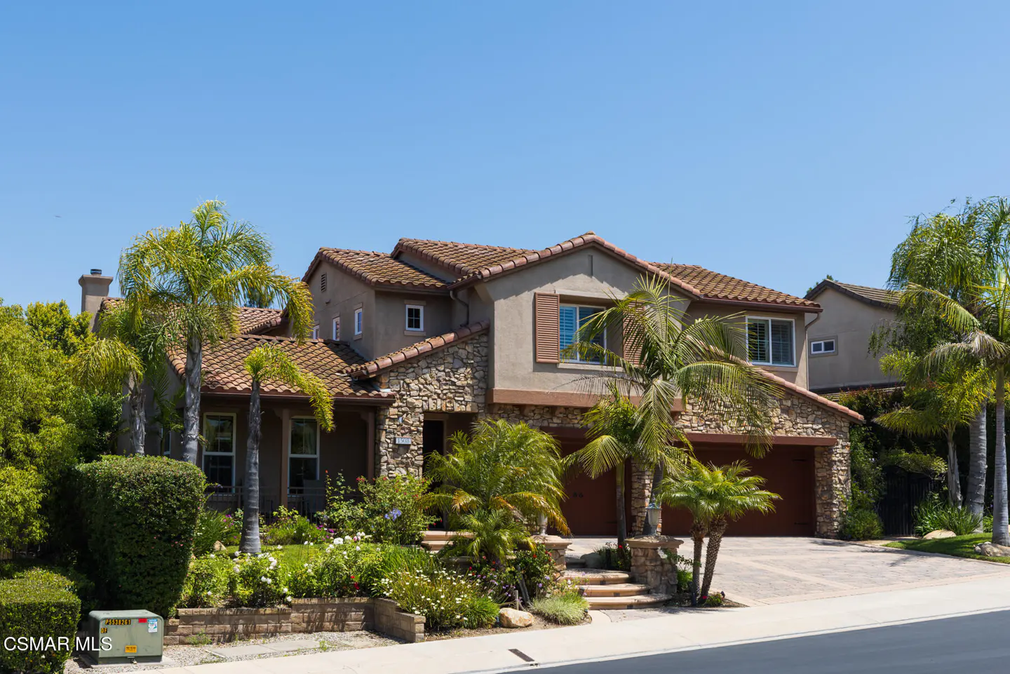 Two-story suburban home with tan stucco, stone facade, tile roof, palm trees, and a paved driveway under a clear blue sky.