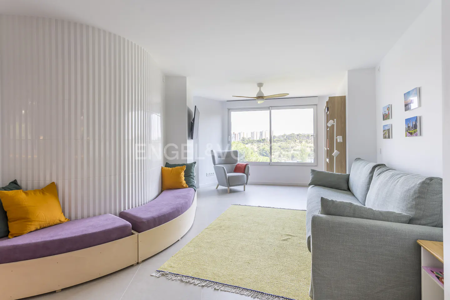 Bright living room with a curved white wall, purple bench with yellow pillows, gray sofa, and a large window with a view.