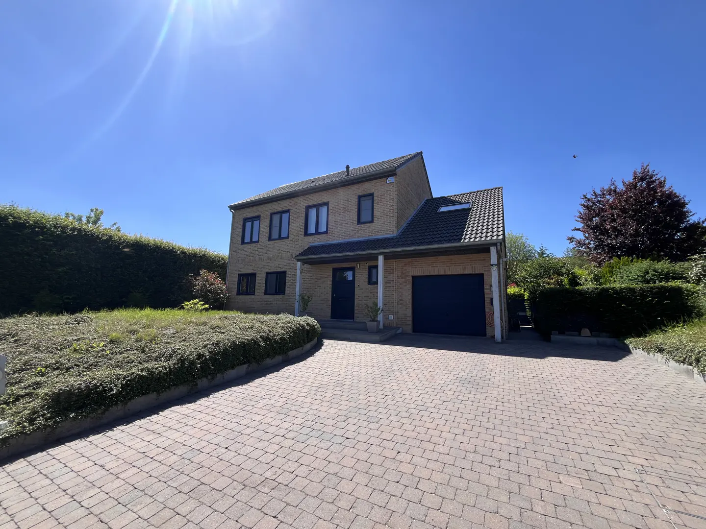 Two-story beige brick house with a dark blue door and garage, a brick driveway, and green bushes under a blue sky.