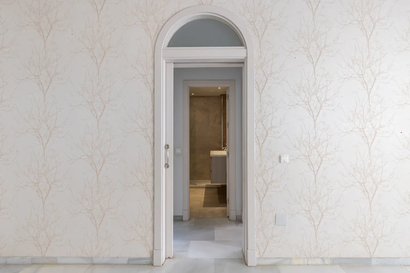 Arched doorway leads to a bathroom. Walls have tree-branch wallpaper. Marble floor.