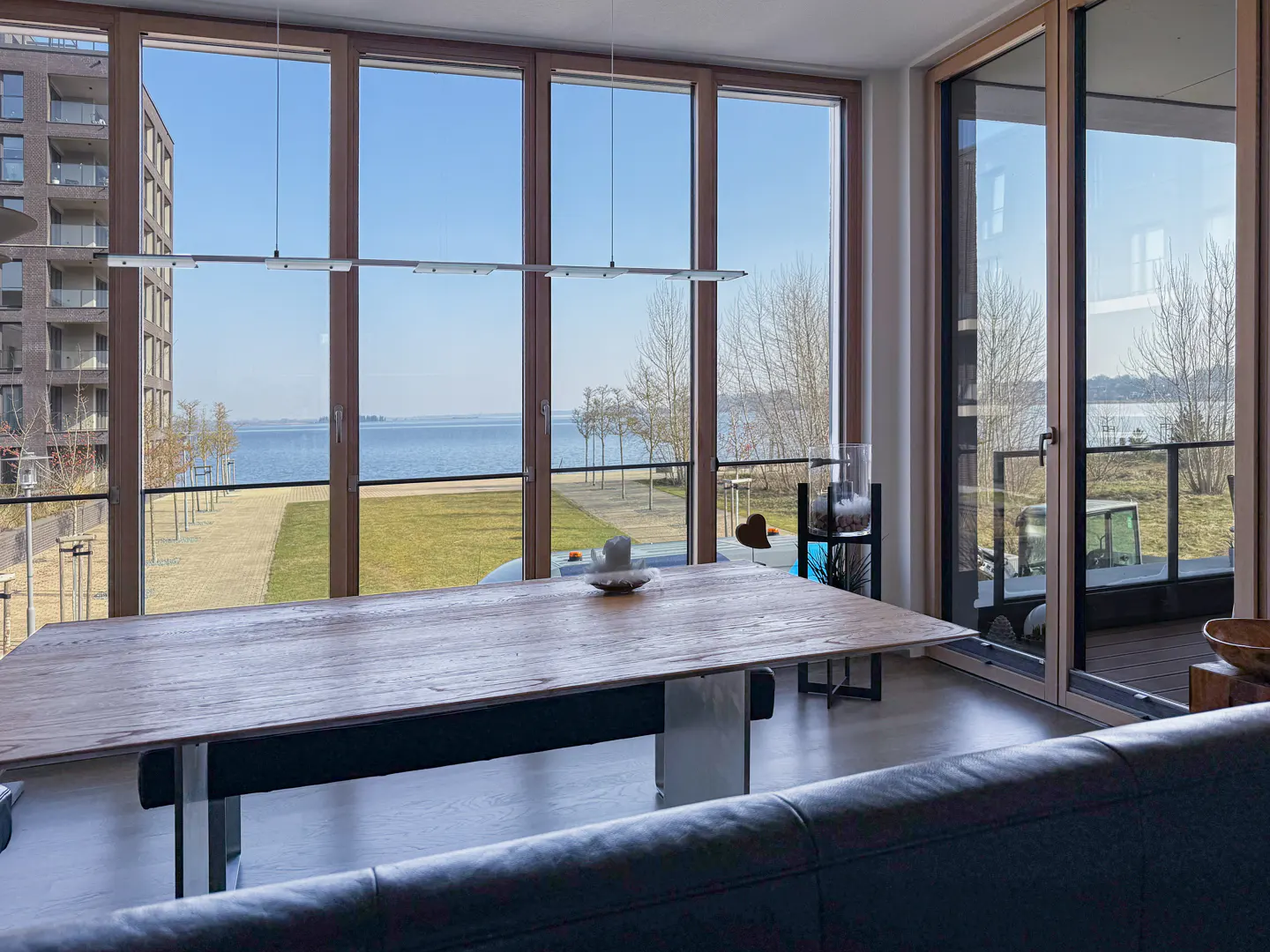 Living room with a large wooden table, floor-to-ceiling windows overlooking a lake, and a black leather couch in the foreground.