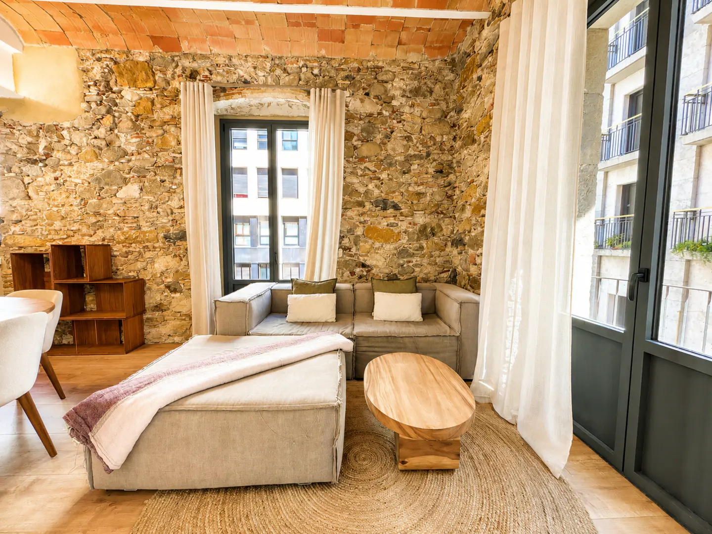 Living room with stone wall, brick ceiling, and wood floors. A gray sofa, ottoman, and wood table sit on a round jute rug. White curtains frame a black window.