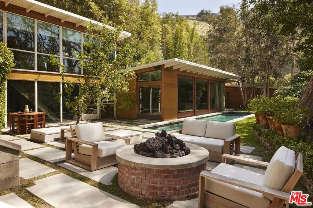 Outdoor patio with a brick fire pit, modern wood furniture, and a pool house with a pool in a lush green backyard.
