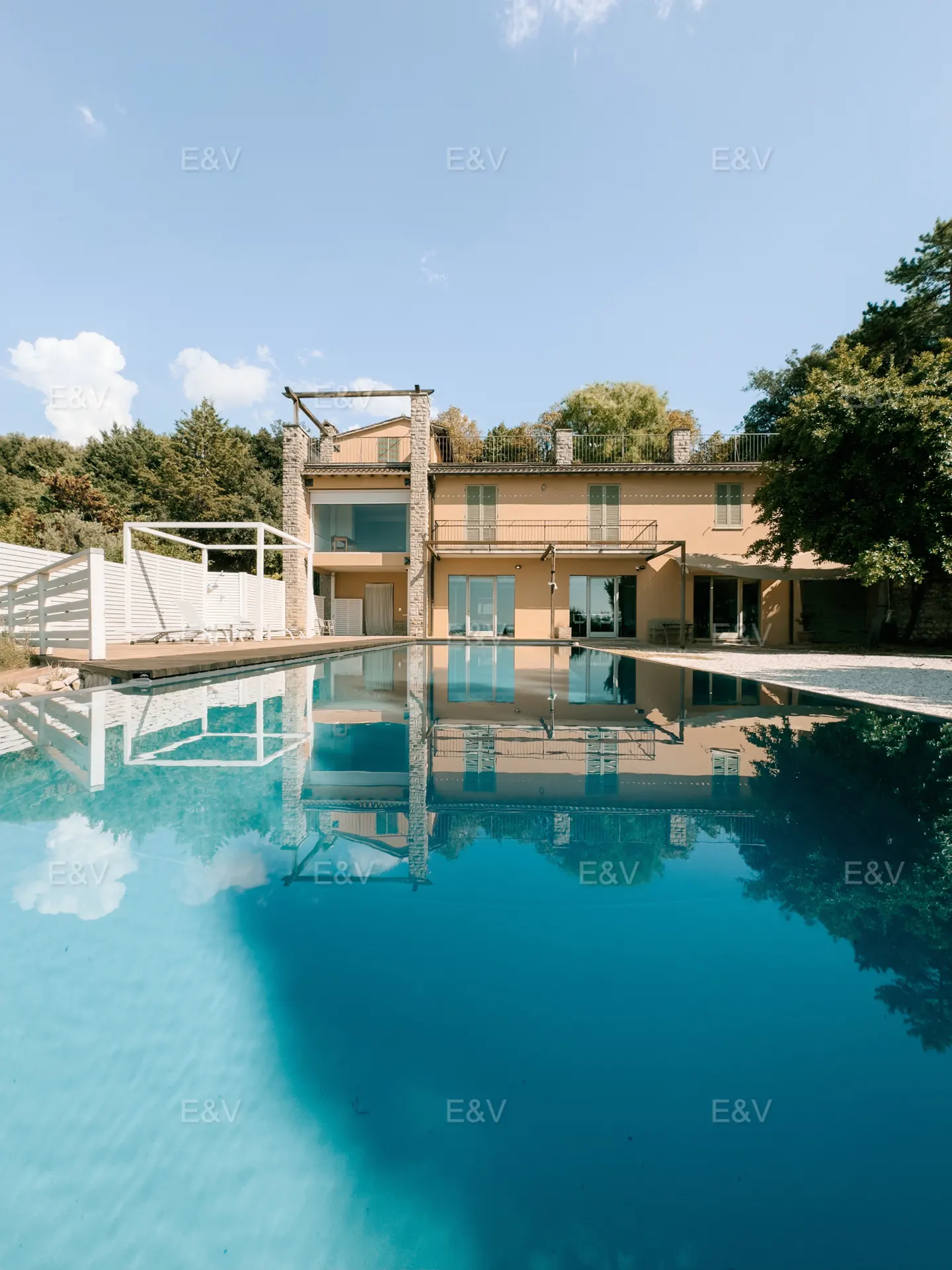 A tan two-story house with a blue pool in front, reflecting the house and sky. Trees are in the background.