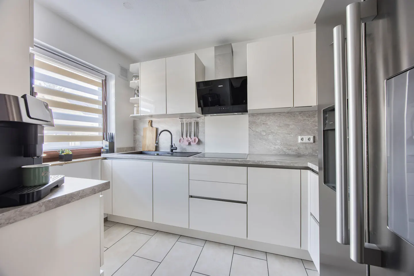 Bright kitchen with white cabinets, gray countertops, and stainless steel refrigerator. A coffee maker sits near a window with blinds.