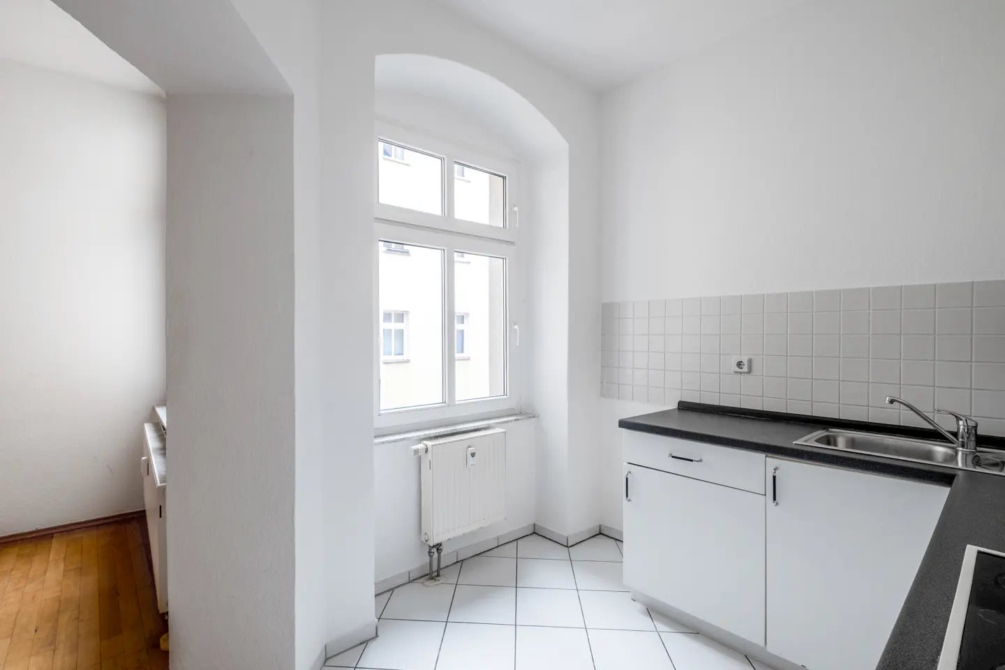 Bright, white kitchen with black countertops, white cabinets, and a stainless steel sink. A window with a radiator sits to the left.