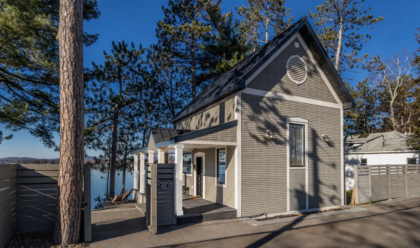 Exterior view of a gray cottage-style house with a black roof, white trim, and a covered porch, surrounded by trees and a lake.
