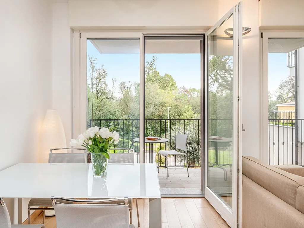 Bright dining room with white table, chairs, and flowers. Open sliding doors lead to a balcony with green trees in the background.