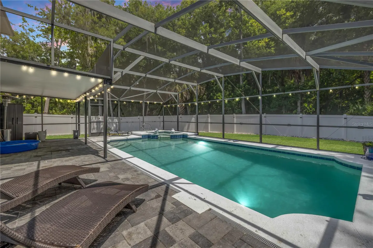 Screened-in backyard pool with a spa, stone patio, two lounge chairs, and string lights under a sunset sky.