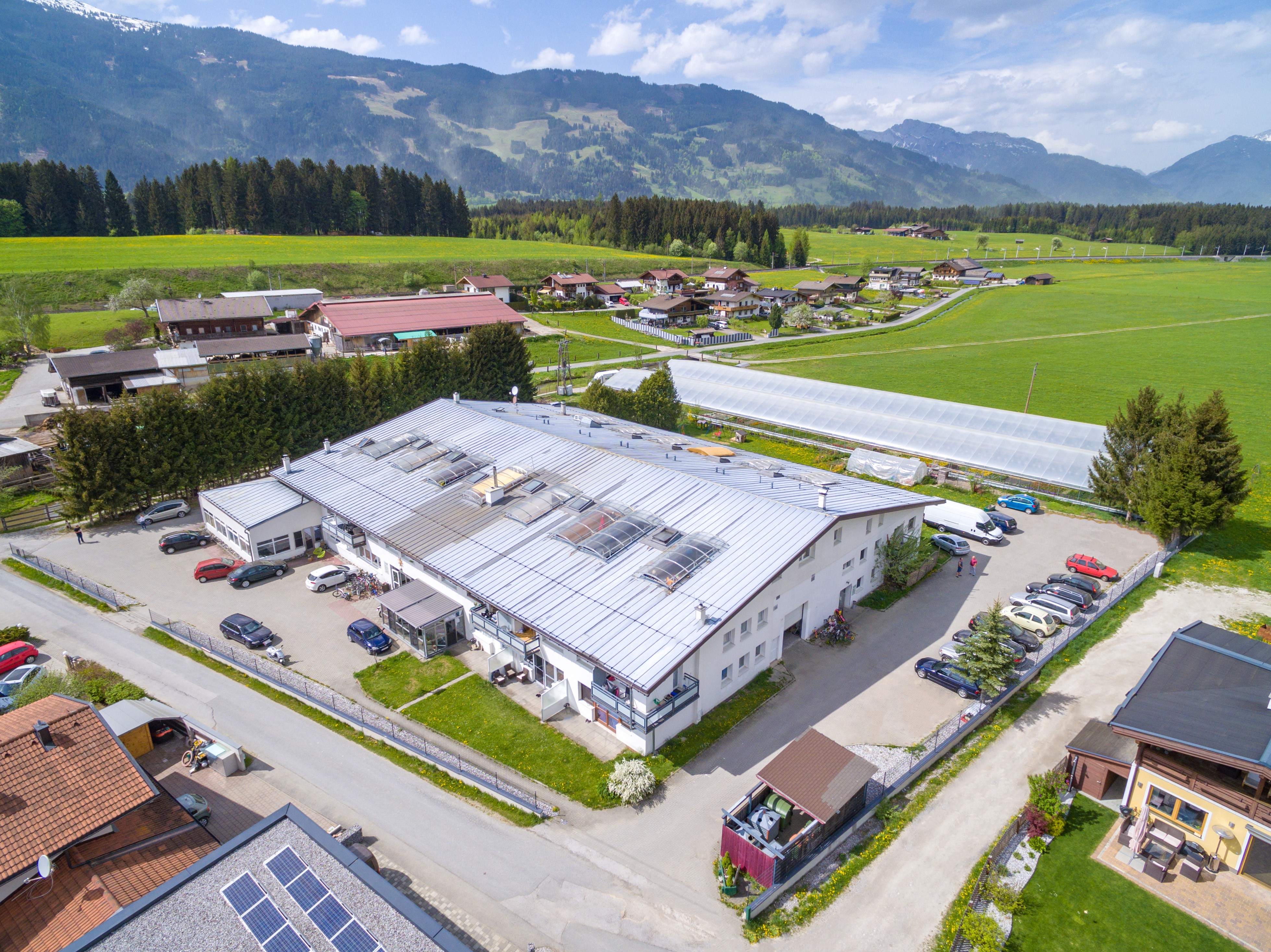 Aerial view of a large, white commercial building with a silver roof, surrounded by green fields, trees, and mountains in the background. Cars are parked in the lot.