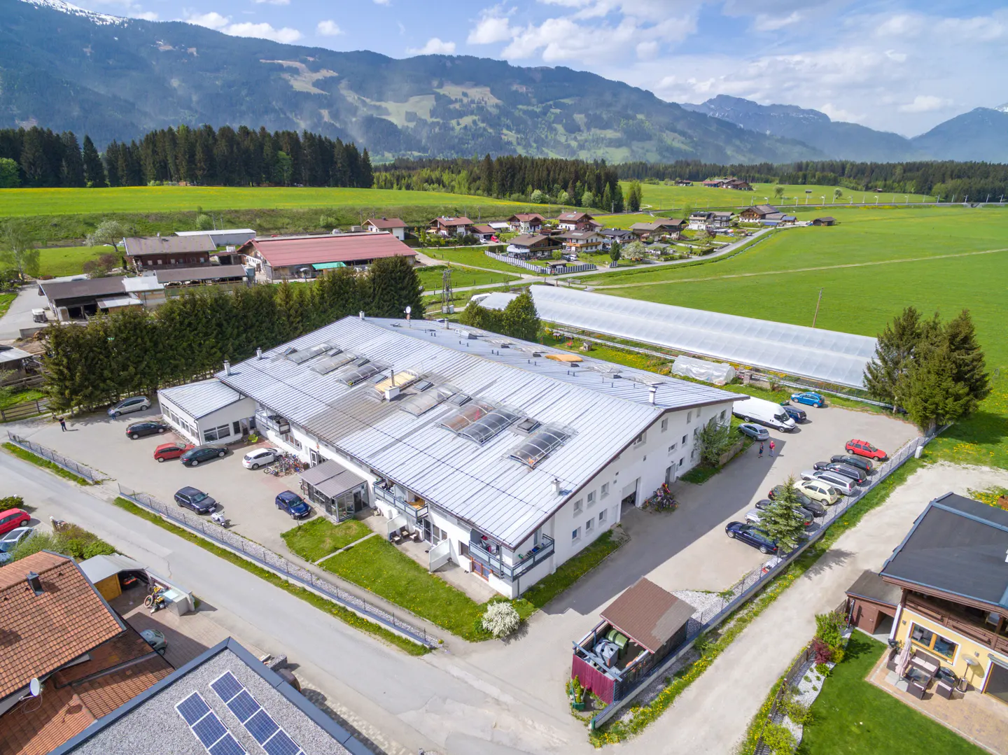 Aerial view of a large, white commercial building with a silver roof, surrounded by green fields, trees, and mountains in the background. Cars are parked in the lot.