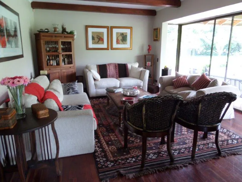 Living room with white sofas, wooden furniture, and a patterned rug on a hardwood floor. Large windows offer a view of greenery.