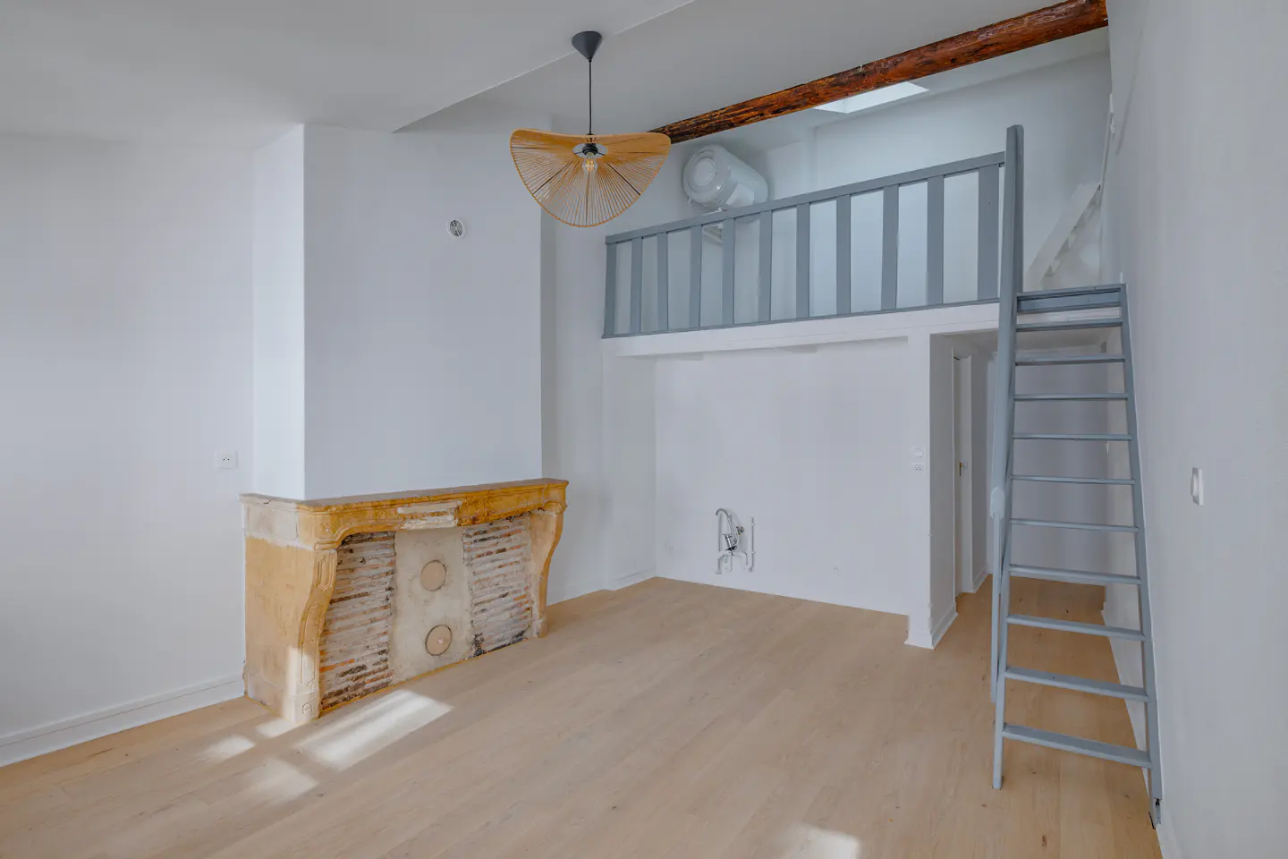 Bright, empty room with light wood floors, white walls, and a gray ladder leading to a loft. A decorative fireplace and modern light fixture add character.