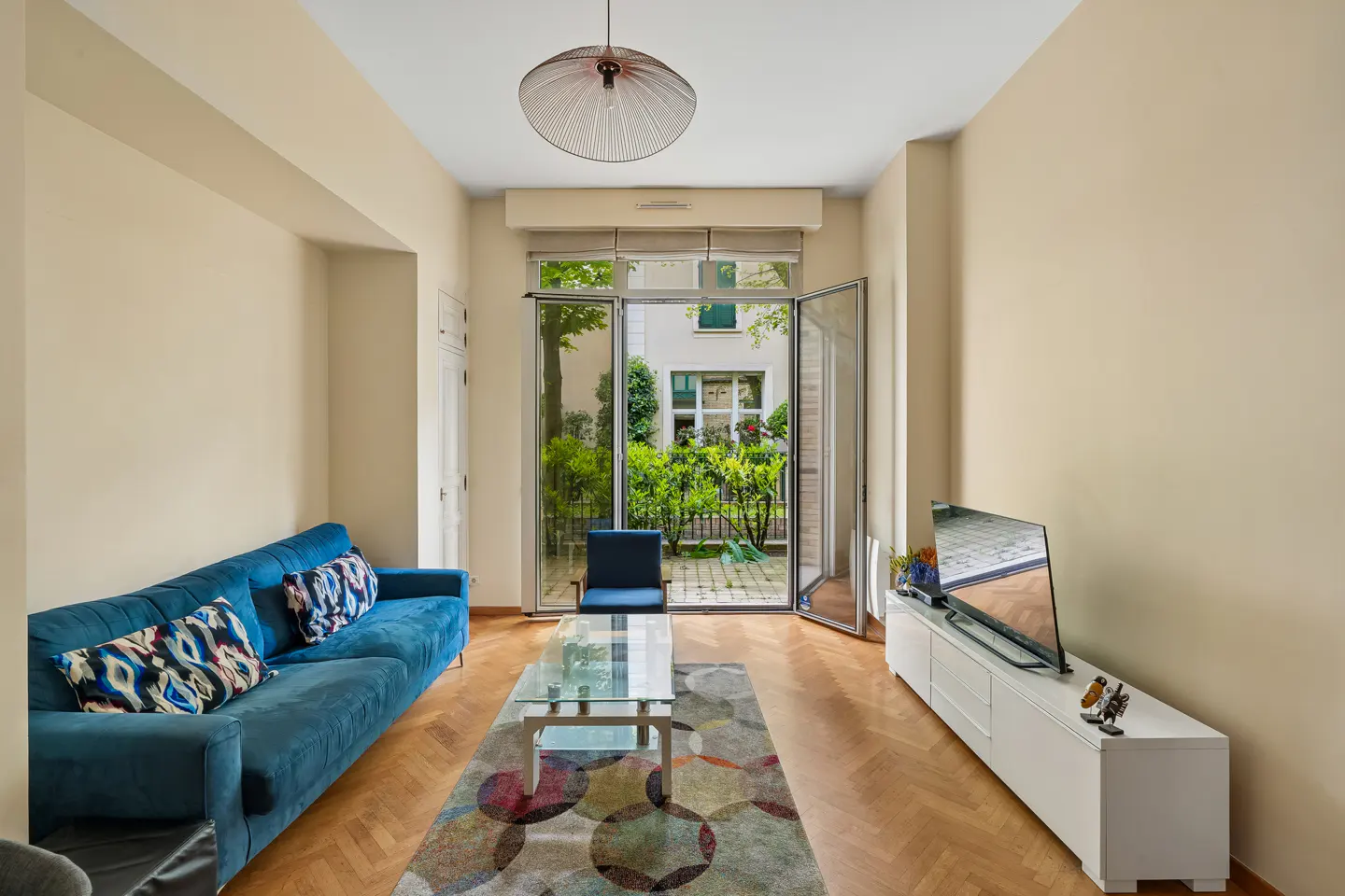 Living room with blue sofa, glass table, and TV. French doors open to a garden view. Neutral walls and wood floors.