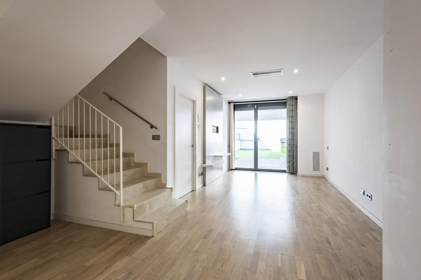 Bright, modern living room with wood floors, white walls, and a staircase. Sliding glass doors lead to a green lawn.