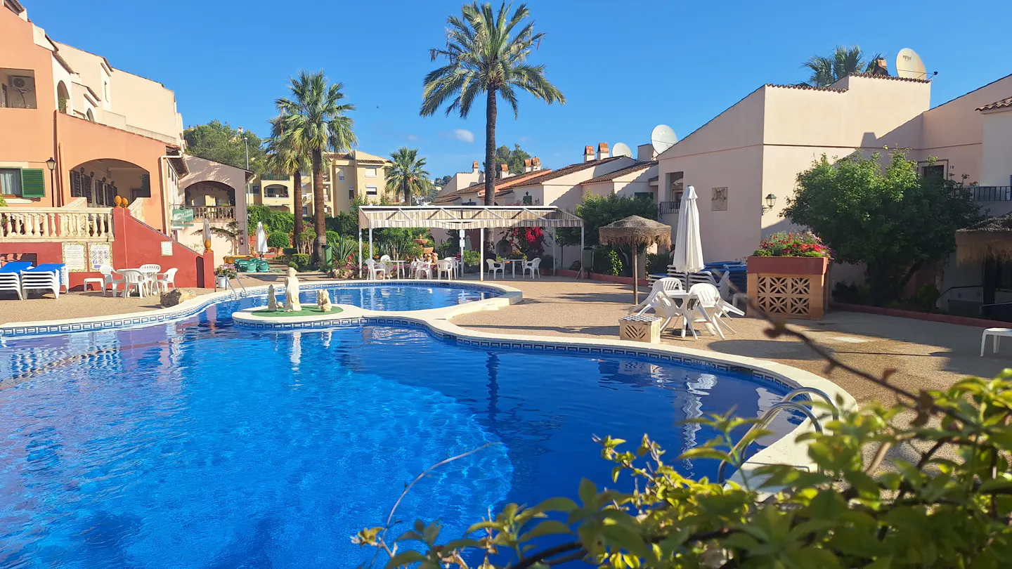 Bright blue pool surrounded by peach buildings, palm trees, and patio furniture under a clear blue sky.