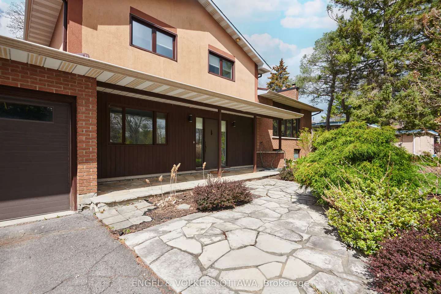 Exterior view of a two-story house with a brown garage door, stone walkway, and green bushes. The house has a tan stucco upper level and a brown wood paneled lower level.