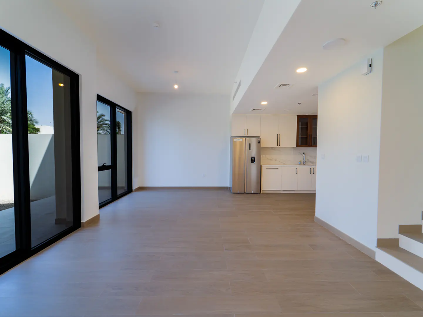 Bright, empty living room with wood floors, white walls, and black-framed windows. A kitchen with stainless steel appliances is visible in the background.
