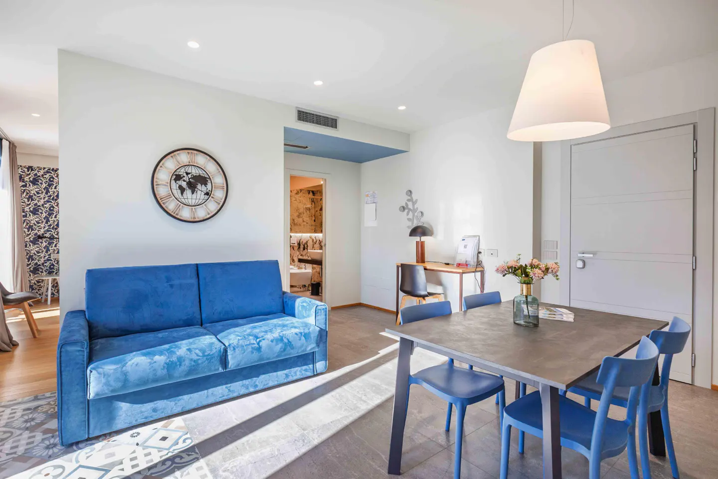 Bright living space with a blue sofa, dining table with blue chairs, and a desk. A world clock hangs on the white wall.