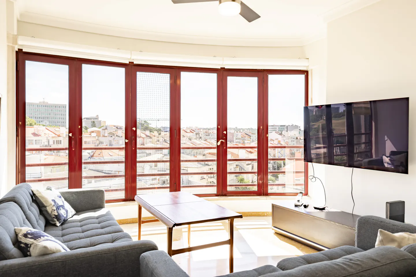 Bright living room with grey sofas, a wooden coffee table, and a wall-mounted TV, facing large windows with a city view.