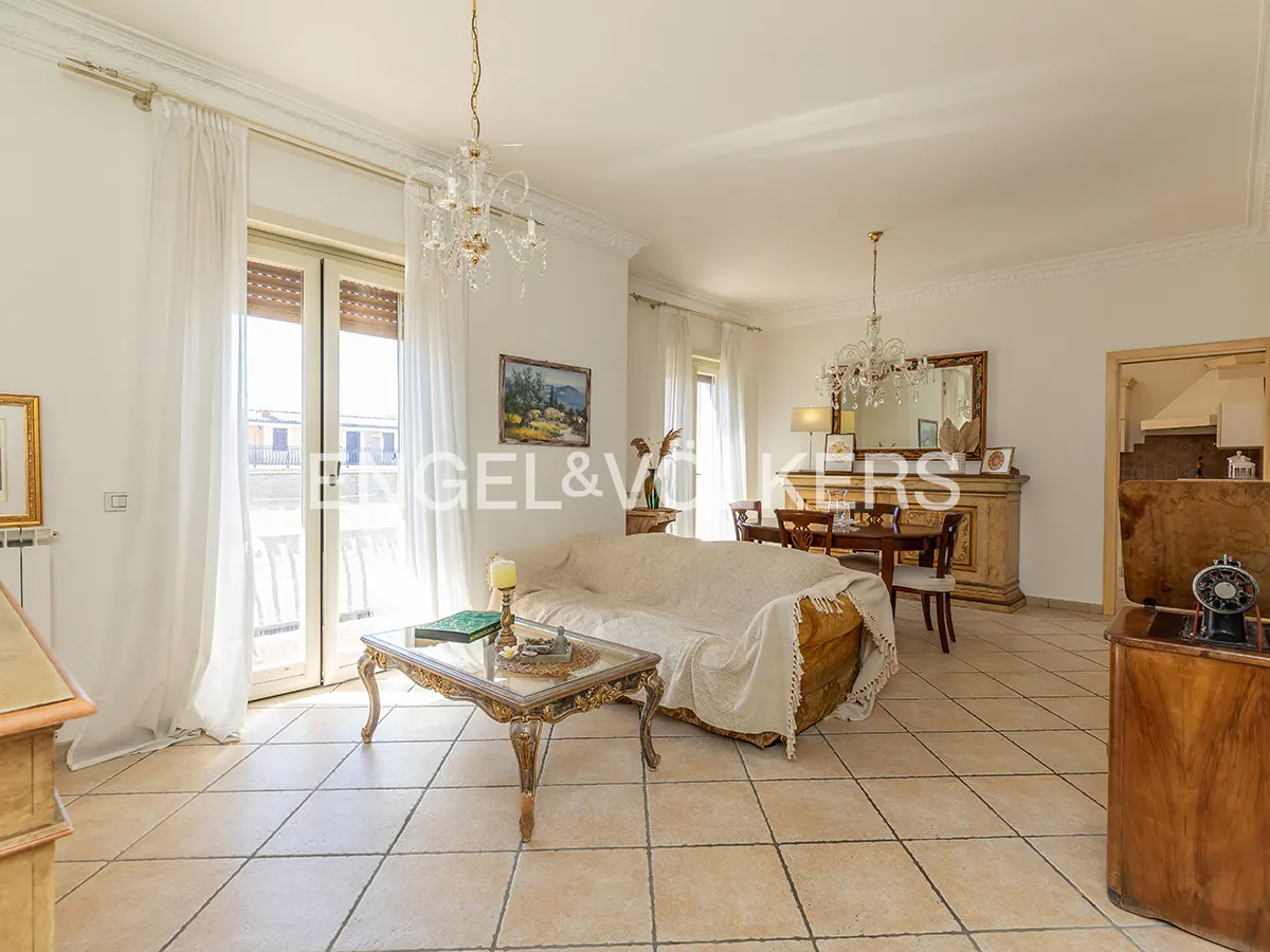 Bright living room with beige tile floors, a sofa covered in a white sheet, and a glass-topped coffee table. Chandeliers hang from the ceiling.