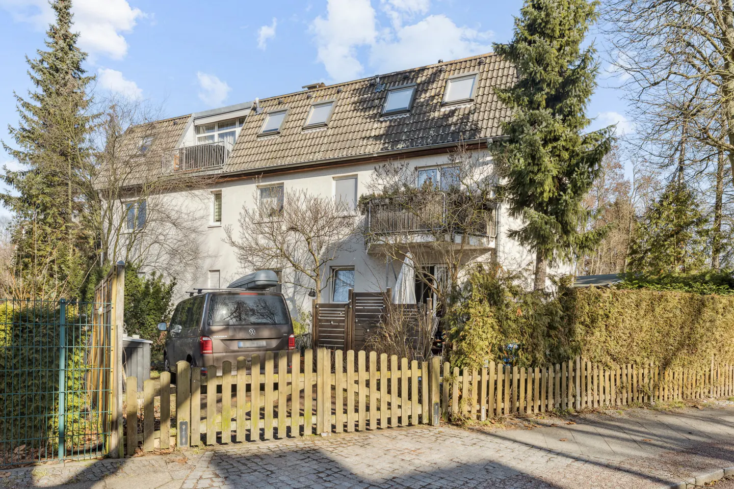 A white multi-story house with a tiled roof, balcony, and a dark van parked behind a wooden picket fence on a sunny day.