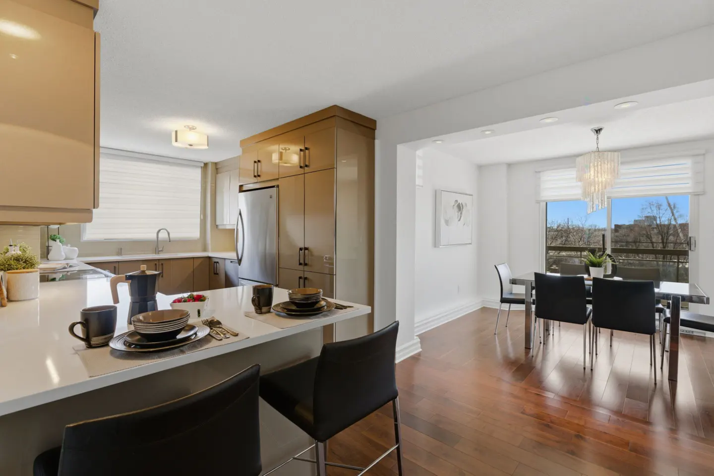 Open concept kitchen and dining area with hardwood floors. Kitchen island with bar stools and place settings. Stainless steel refrigerator. Chandelier over dining table.