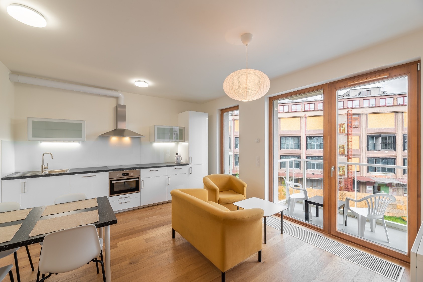 Modern kitchen with white cabinets, yellow armchairs, dining table, and a balcony overlooking a city building. [AI-generated text]