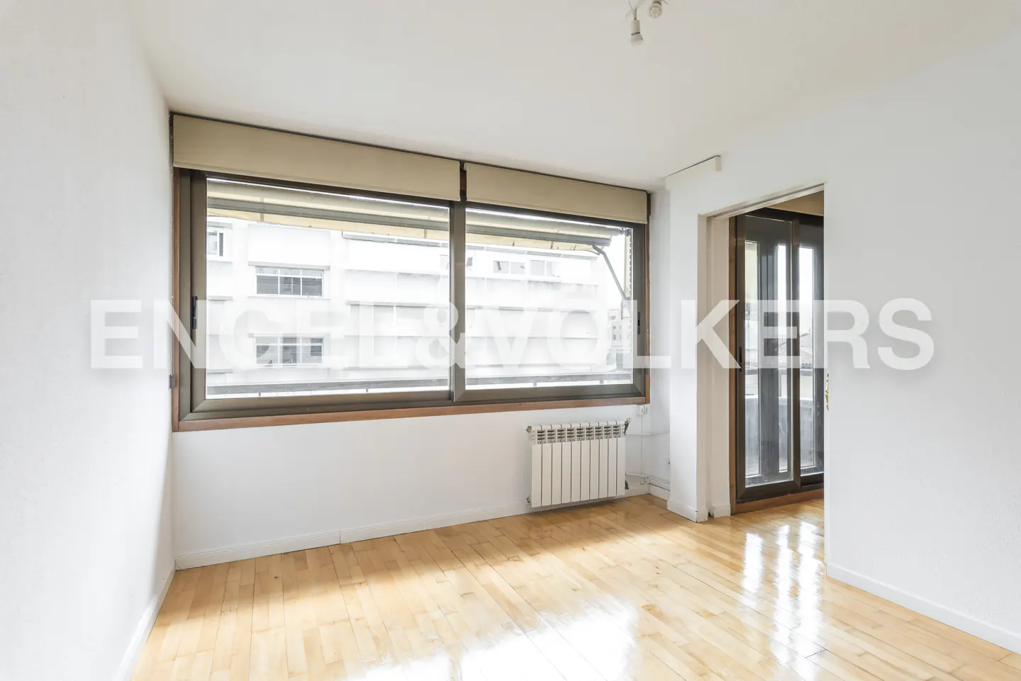 Empty room with white walls, light wood floors, and a large window with blinds. A radiator sits below the window. Doorway to another room is visible.