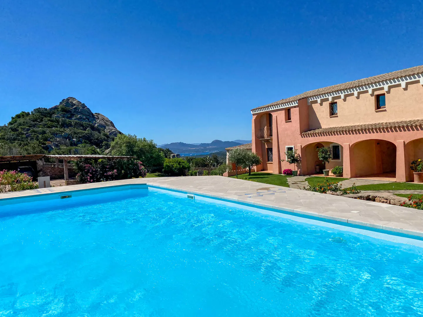 A bright blue swimming pool in front of a peach-colored Mediterranean villa with a mountain and sea view under a clear sky.